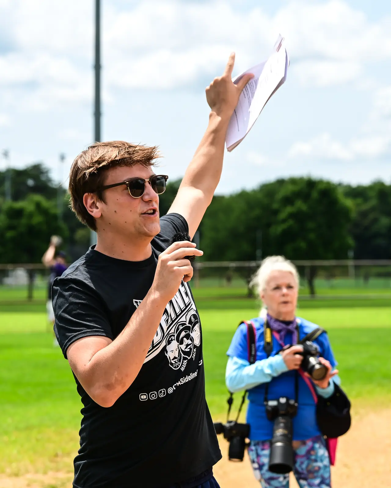 Field announcer raising a sheet of paper while addressing the Summer Slugfest crowd.