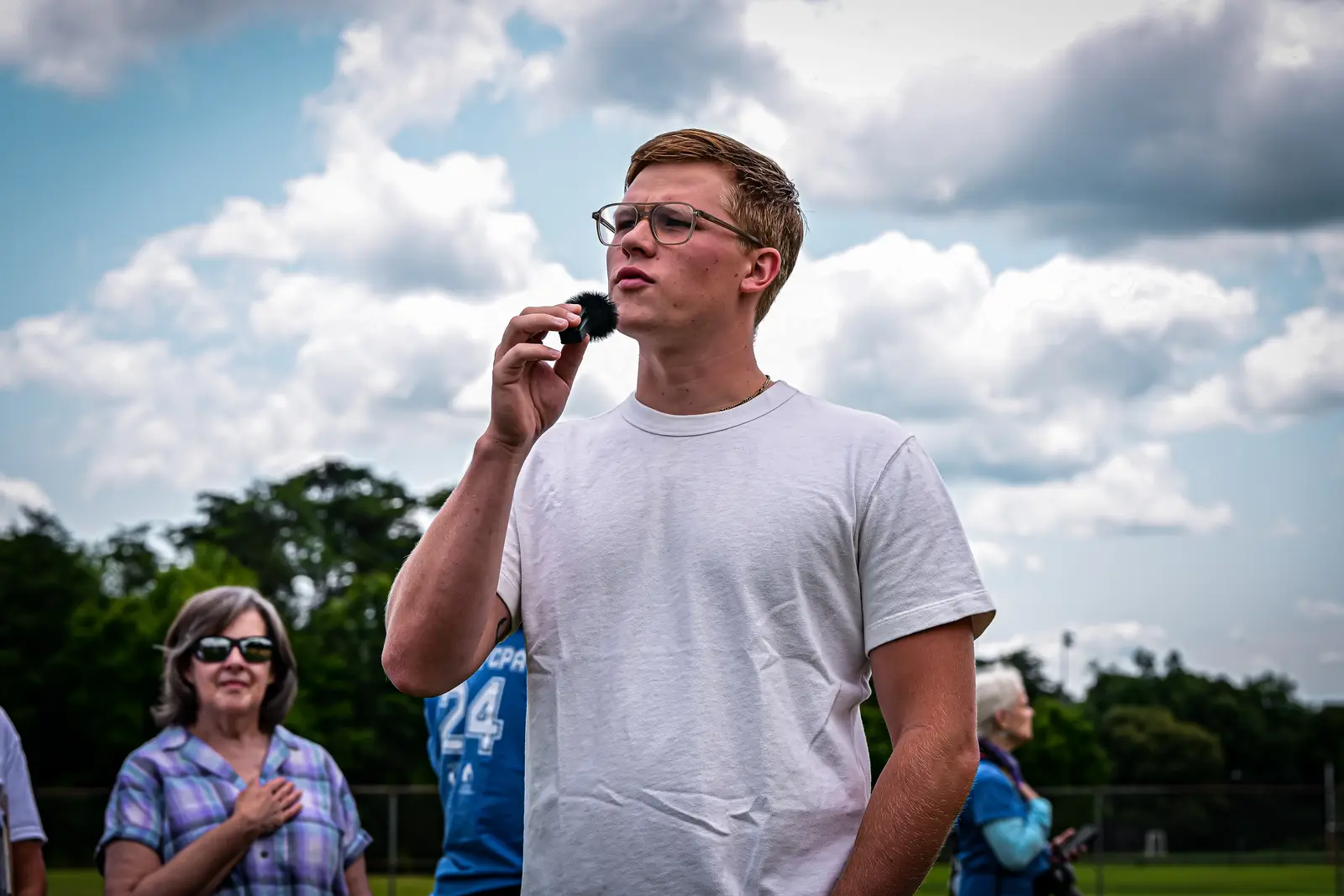 Summer Slugfest field announcer speaking into a handheld microphone beneath a cloudy sky.
