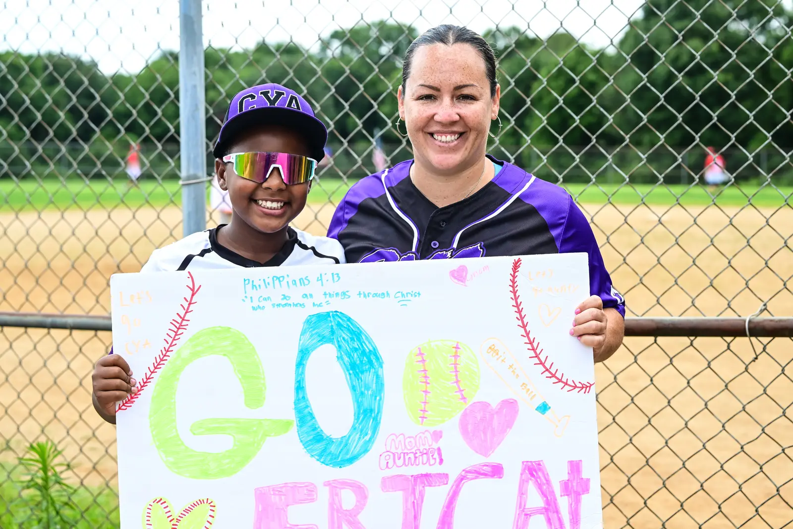 Two CYA supporters holding a colorful handmade sign beside the softball field fence.