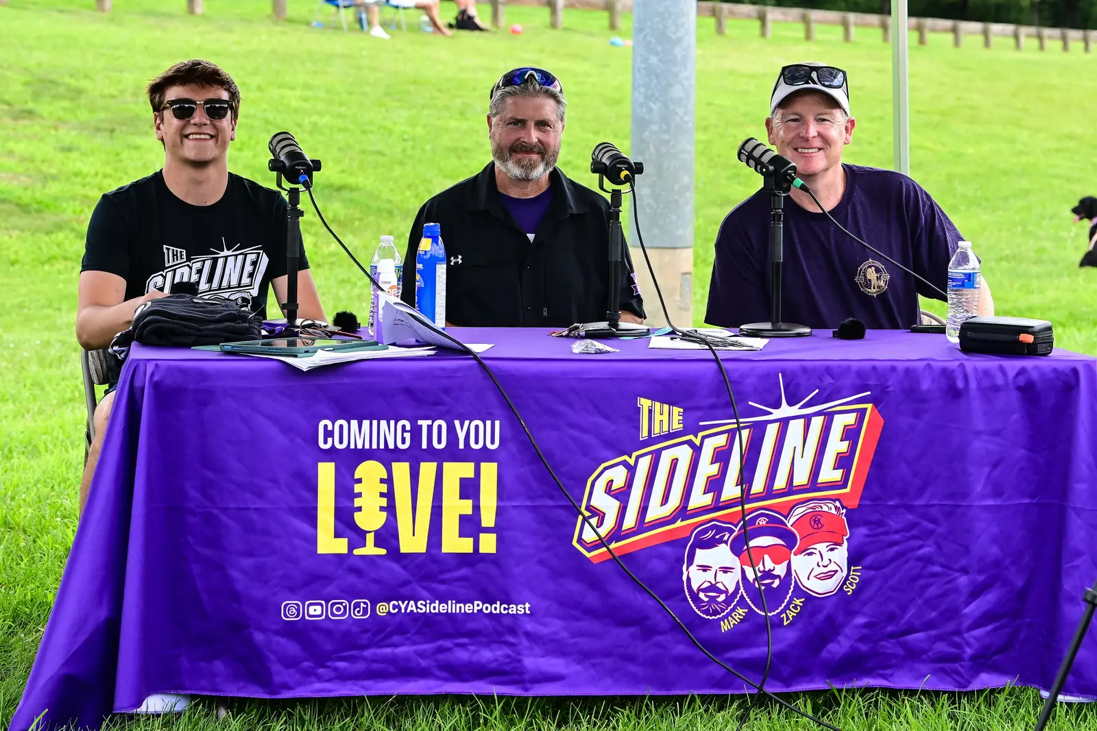 Three Sideline Podcast hosts seated behind a purple broadcast table at Summer Slugfest.